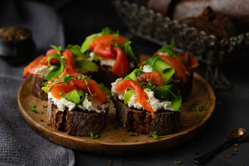 Homemade sandwich with rye bread, salmon, avocado, cottage cheese and microgreen sprouts on wooden plate on dark background. Tasty breakfast close-up