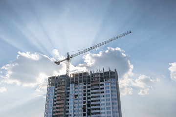 construction crane above a residential block of flats under construction against the blue sky and the sun's rays