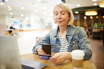 Content attractive mature woman sitting in cafe and using smartphone while scanning credit card for online payment, online shopping concept