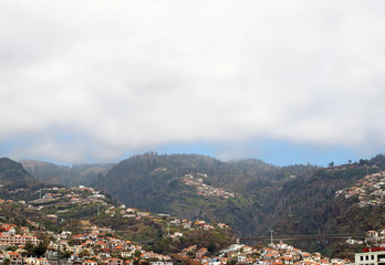 Fototapeta na wymiar an aerial panoramic cityscape view of the city of funchal in madeira with houses in front of tree covered mountains and white clouds