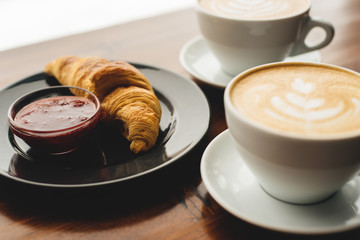 Two cups of cappuccino with latte art and croissant with jam on the wooden table