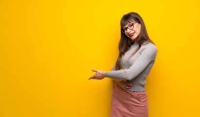 Woman with glasses over yellow wall pointing back and presenting a product