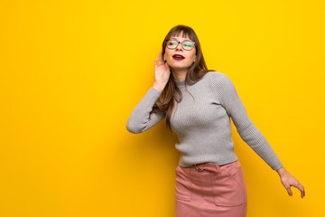 Woman with glasses over yellow wall listening to something by putting hand on the ear