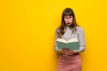 Woman with glasses over yellow wall surprised while enjoying reading a book