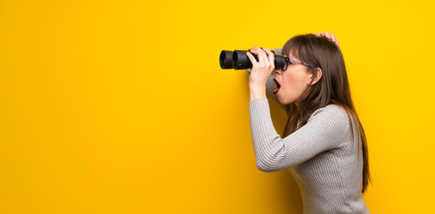Woman with glasses over yellow wall and looking in the distance with binoculars