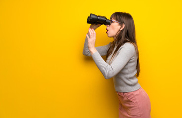 Woman with glasses over yellow wall and looking in the distance with binoculars