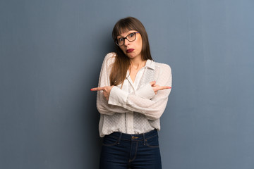 Woman with glasses over blue wall pointing to the laterals having doubts