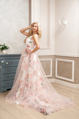 Beautiful blonde woman in pastel floral long puffy dress with makeup and long curly hairstyle...