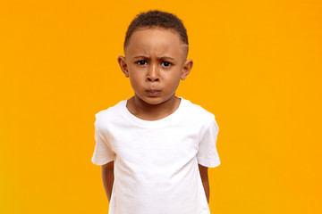 Studio image of displeased frowning Afro American little boy in white t-shirt having grumpy...