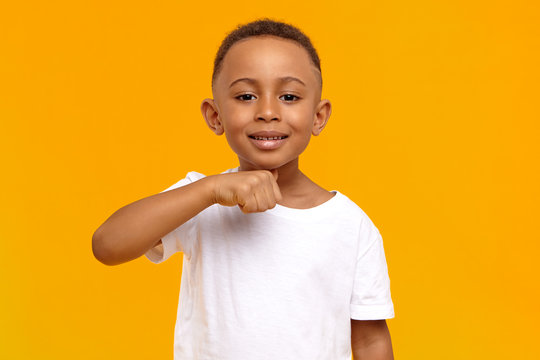 I will cut your head off. Picture of positive cheerful dark skinned schoolboy having fun in yellow studio, making joke, holding thumb at his neck, showing menacing warning gesture, smiling broadly