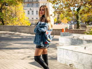 cute blonde girl with flowing hair in a jeans jacket standing on the street against the background of yellow trees