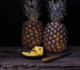 Sliced pineapple on old rustic desk with knife and with two pineapples behind. Black background.