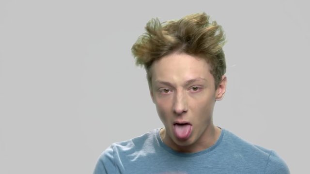 Rude teen boy making face grimace. Teenage boy sticking his tongue out against gray background. Adolescent guy teasing at camera.