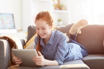Portrait of smiling teenage girl using digital tablet lying on sofa at home lit by sunlight, copy...