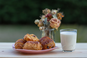 Buns with raisins and french croissant. A glass of milk on rustic table. Dinner in an orchard in summertime. Bouquet of flowers. Dessert. Soft focus. Summer twilight. Blurry background. Copy space.