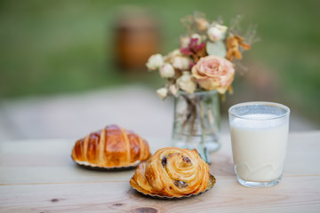 French croissant and buns with raisins, a glass of milk on a wooden table. Dinner in an orchard in summertime. Bouquet of flowers. Dessert. Soft focus. Nature wallpaper blurry background. Copy space.