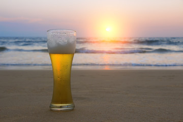 Misted glass of cold Chang beer on the sand at the background of seascape, sunset sky and sea waves.