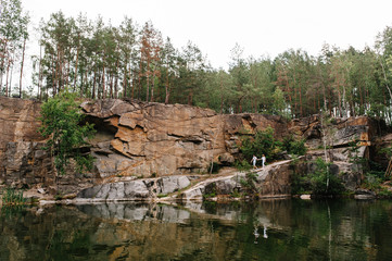 Fototapeta na wymiar Reflection of couple people going near lake on background of rocks. Landscape of an old flooded industrial granite quarry filled with water. Canyon. Place for text and design. full length
