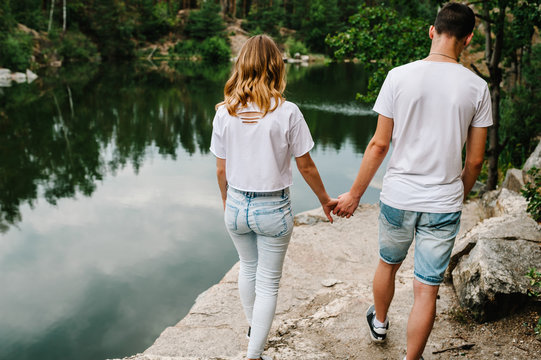 Legs Young Couple Going On Stone Near Lake. Full Length. Man's And Woman's View From The Back On Background Of Rocks. Landscape Of An Old Industrial Granite Quarry. Close Up. People Are Holding Hands