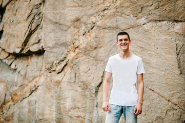 A man makes a grimace, stand on background of the large rocks wall on a big stone. upper half. looking at the camera. Close up. Place for text and design. Man poses at cliff of mountain on nature.