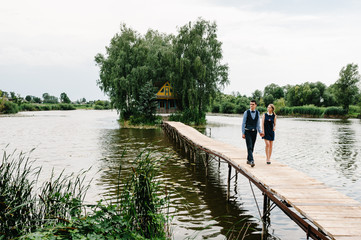 The young couple is holding hands, going on a wooden bridge above the lake on background a old house on an island near of trees. Nature, landscape. full length.
