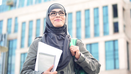 Inspired young female student wearing hijab smiling, standing outdoors on campus