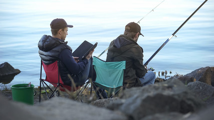 Bored men catching fish, using tablet to search tutorials on fishing websites