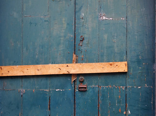 close up of an old peeling green painted wooden plank door barred shut with a piece of timber and rusty nails with a lock and handle