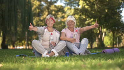 Two women showing thumbs up sitting on grass after doing exercises, positive
