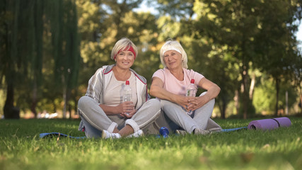 Fototapeta premium Two women sitting on grass and smiling into camera after finishing doing yoga