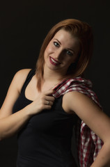 Happy young woman on the black background. Woman is holding a checkered shirt over her left shoulder. Vertically. 