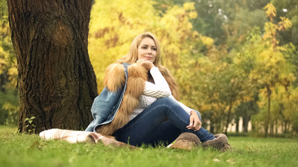 Pretty woman sitting on plaid under tree in autumn park, posing for camera