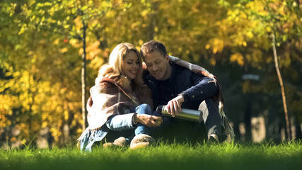 Man pouring hot tea from thermos, loving couple covered with plaid having picnic