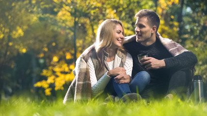 Couple covered with plaid sitting in autumn park, drinking hot tea and talking
