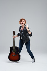 Young woman musician in a black leather jacket with an acoustic guitar in hand on a gray background. He laughs and plays rock and roll loudly. Full-length portrait