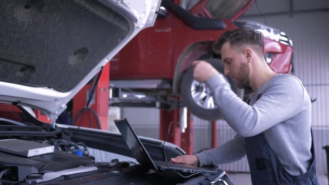 Modern Computer Technology In Car Repair, Professional Technician Male Uses Laptop For Diagnostics Vehicle With Open Hood At Service Station