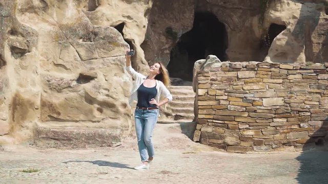 A young blond woman with a smiling face in casual comfortable clothes makes a photo against the background of a historic landmark. Background of the cave of the ancient city Uplistsche.