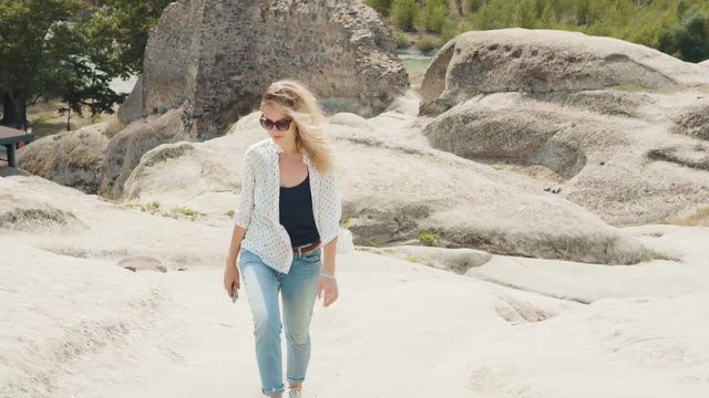 A young blonde woman in jeans and a white shirt and sunglasses walks near of the ancient city carved into the rocks. Tourist in picturesque historical Georgia. Bright, positive sunny day