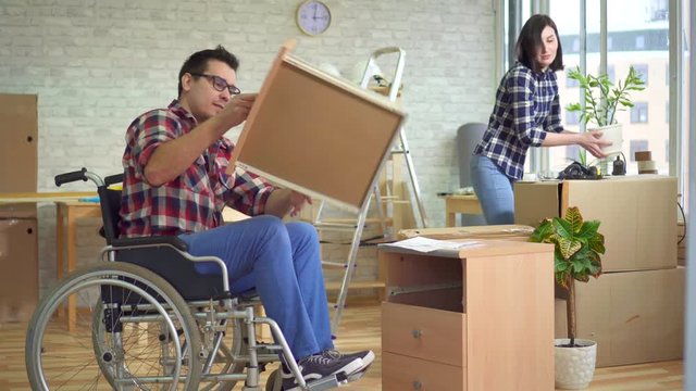 Concept of moving, a disabled man in a wheelchair collects a bedside table next to his wife