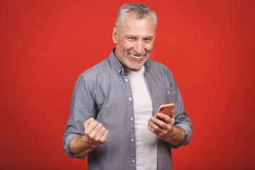 Happy mature senior man in casual using smartphone and making winner gesture isolated over red background. Using phone.
