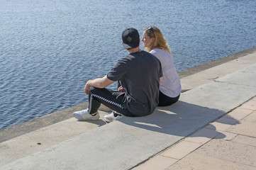 Young couple sit in sunshine watch water glitter on stone pier