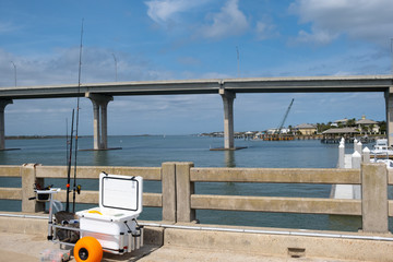 Fishing poles and gear set-up on a pier overlooking the water with a bridge in the backround under a clear blue sky