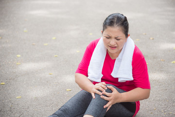Asian aged woman suffering with knee ankle pain while running in park. Middle aged female sitting on the ground and holding painful ankle.