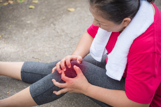 Asian Aged Woman Suffering With Knee Ankle Pain While Running In Park. Middle Aged Female Sitting On The Ground And Holding Painful Ankle.