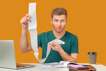 The indignant man holds checks, not understand from where the big sums, not ready to unforeseen expenses. Isolated on an orange background in the Studio