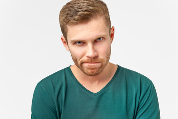 Puzzled man keeps lips pressed, has frustrated facial expression, frowns face in displeasure, being deep in thoughts. Male dressed in casual outfit, models over white wall