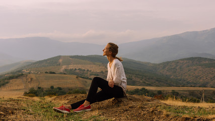 Naklejka na ściany i meble Profile portrait of young pretty girl who is smiling, wearing casual outfit, enjoying, sitting on the yellow grass in mountains, foggy sky on the background