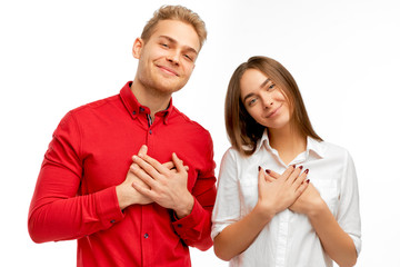 A good-natured attractive young people has a friendly expression, holds hands on heart. The young friends shows love and great sympathy, isolated by white wall in the Studio