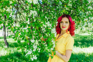 Beautiful and young woman with bright red hair and ref lips standing next to a blooming apple tree in a yellow dress.