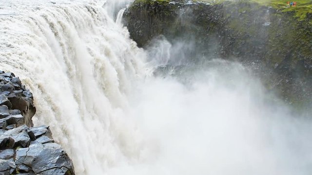 Icelandic Dettifoss waterfall closeup in Iceland gray water mist in slow motion and rocky cliff spraying and people standing far on overlook